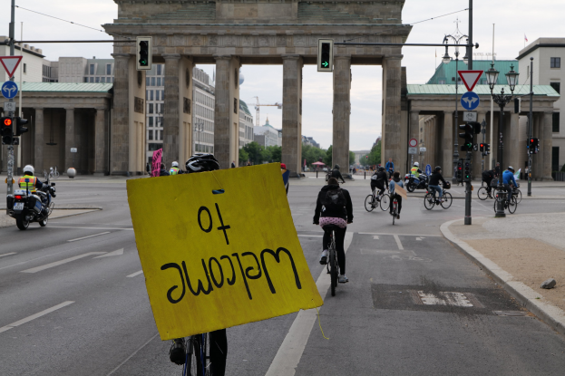 Eine Gruppe von Radfahrern fährt am Brandenburger Tor in Berlin vorbei, einer hält ein gelbes Schild, mit Laternen, Verkehrszeichen, Gebäuden, Bäumen und einem klaren Himmel im Hintergrund.