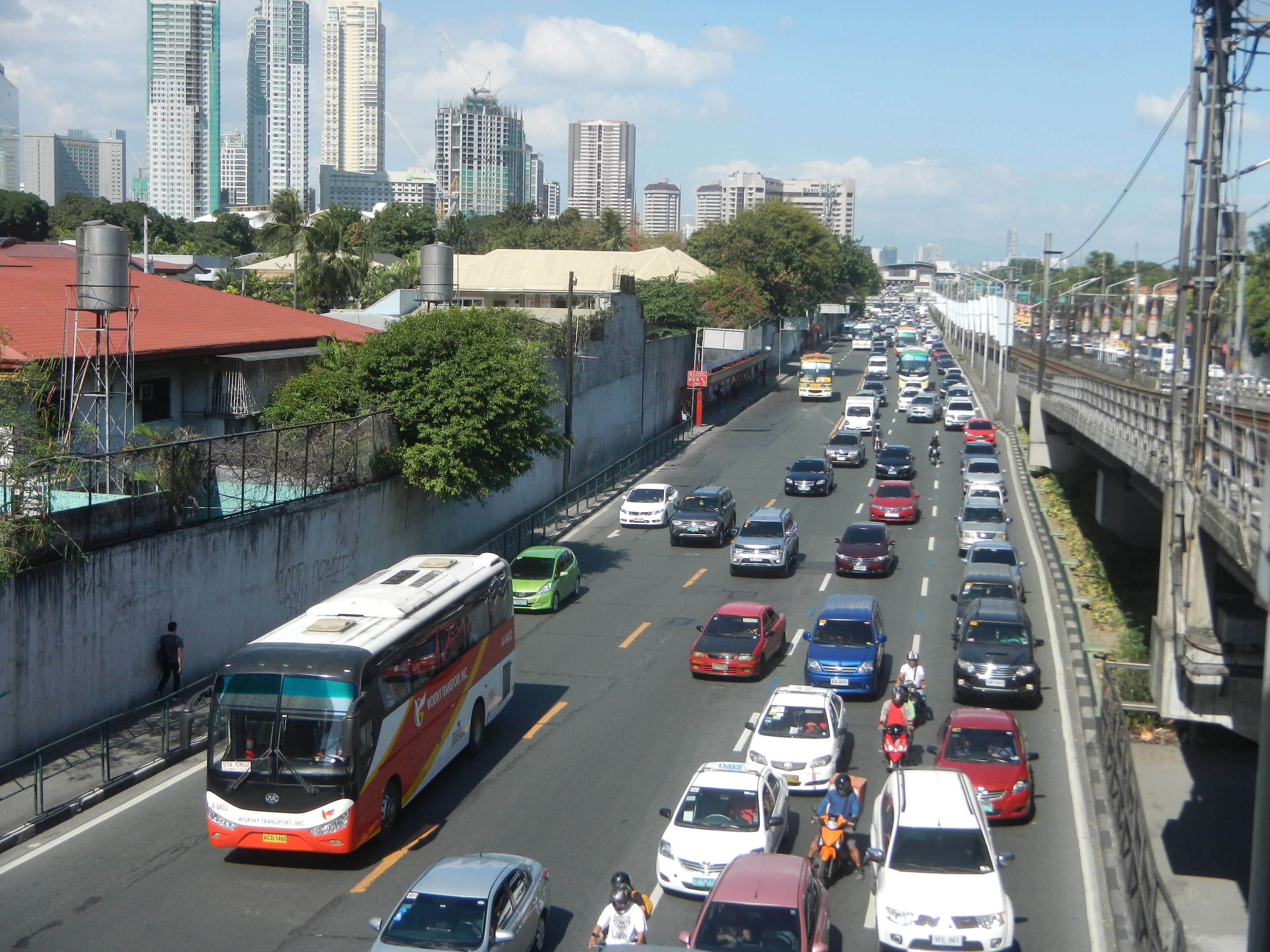 Voller Stadtverkehr mit Autos, Bussen und Motorrädern auf einer Straße mit Strommasten und -leitungen auf der rechten Seite und Gebäuden, Bäumen und bewölktem Himmel im Hintergrund.