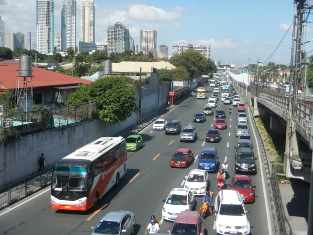 Voller Stadtverkehr mit Autos, Bussen und Motorrädern auf einer Straße mit Strommasten und -leitungen auf der rechten Seite und Gebäuden, Bäumen und bewölktem Himmel im Hintergrund.