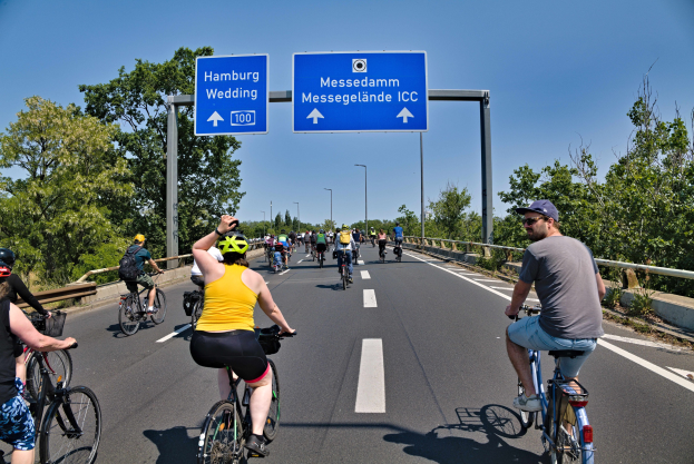 Gruppe von Radfahrern in Helmen auf einer Straße mit Bäumen auf einer Seite und Laternen im Hintergrund unter einem klaren blauen Himmel, mit einer Tafel, die eine Radtour in Hamburg anzeigt.
