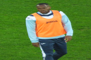 Ein Fußballer in einem orangen T-Shirt und blauen Hosen läuft entschlossen über ein grünes Feld.