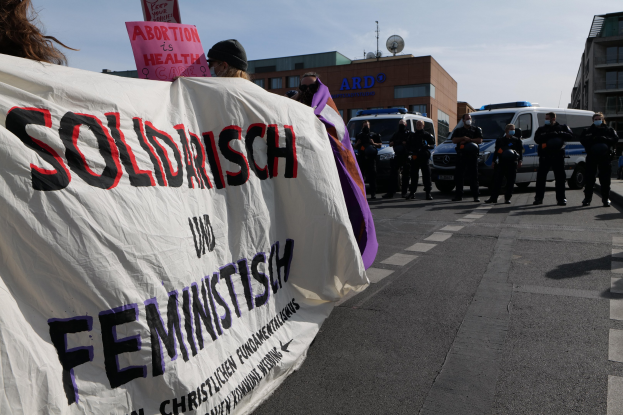 Eine Gruppe von Menschen marschiert mit einem Transparent, auf dem 'Solidarität und Feminismus' steht, eine Straße entlang, mit parkenden Fahrzeugen und Gebäuden im Hintergrund unter einem klaren blauen Himmel.