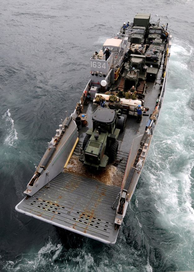 Luftaufnahme eines großen Militärschiffs auf dem Meer mit einem Militärfahrzeug an Bord, umgeben von Menschen und Ausrüstung, mit einem Wasserzeichen unten.