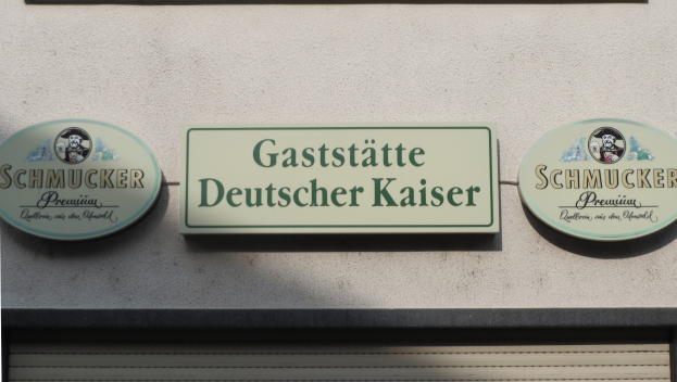 Drei Schilder mit der Aufschrift "Gaststätte Deutscher Kaiser Schmucker" sind an einer Gebäudewand über einer geschlossenen Jalousie angebracht.