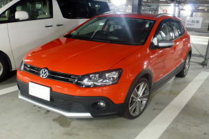 Orange Volkswagen Polo Cross 1.6 TDI Highline DSG in einer Tiefgarage neben einem weißen Lieferwagen geparkt, mit einem Metallzaun, einer Tafel mit Text, Deckenleuchten und einer Dachkonstruktion im Hintergrund.