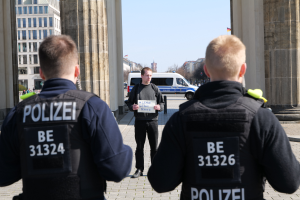 Drei Polizeibeamte stehen vor einem Gebäude, ein Mann in der Mitte hält ein Papier, flankiert von Säulen, mit Fahrzeugen, Menschen, Bäumen, Gebäuden und Himmel im Hintergrund.