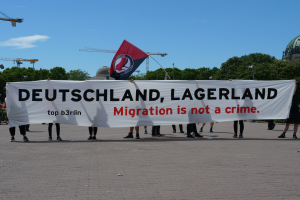 Eine Gruppe von Menschen mit einem Banner, auf dem "Deutschland, Lagerland Migration ist kein Verbrechen" steht und einer Flagge, mit Straßenlaternen, Bäumen, Kränen, einem Gebäude und einem bewölkten Himmel im Hintergrund.