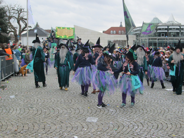 Eine Gruppe von Menschen in Hexenkostümen, die schwarze und lila Kleider und Hüte tragen und die einige Flaggen halten, gehen eine Kopfsteinpflasterstraße entlang, gesäumt von einem Geländer auf der linken Seite und Gebäuden, Bäumen und einem klaren blauen Himmel im Hintergrund.
