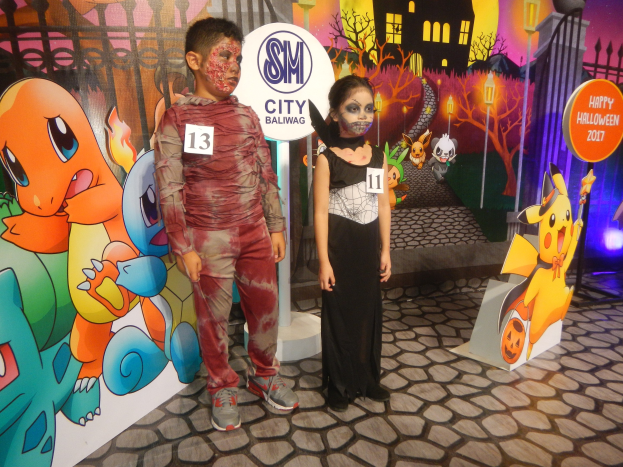 Zwei Kinder in Halloween-Kostümen stehen vor einer bemalten Wand, die eine Straßenszene zeigt, mit Informationsschildern und festlicher Beleuchtung in der Nähe.