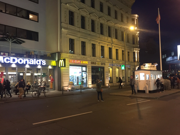 Eine Gruppe von Menschen geht nachts eine Straße in Berlin, Deutschland, entlang mit Gebäuden, Laternen, einer Fahne, Geschäften, Fahrrädern und dem Himmel.