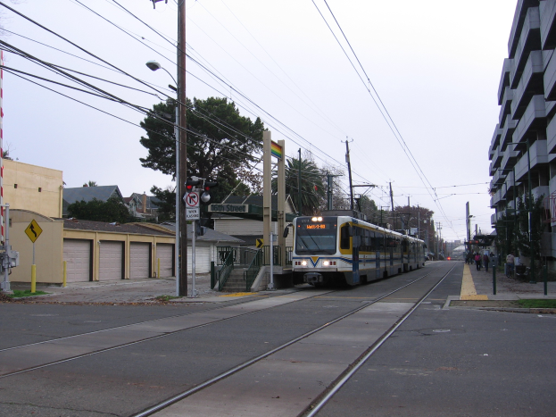 Ein Nahverkehrszug fährt eine Straße entlang, die von hohen Gebäuden, Strommasten und einigen Fußgängern gesäumt ist, mit Bäumen und einem klaren blauen Himmel im Hintergrund.