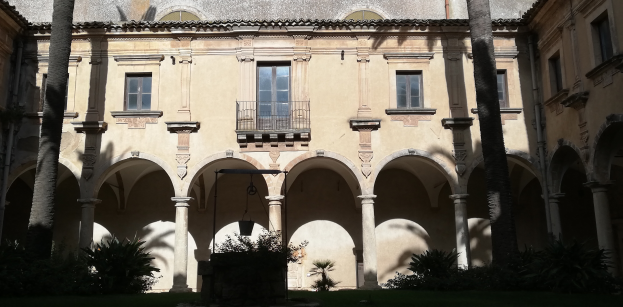 Ein Geb├Ąude mit Fenstern, Gel├Ąndern, Pfeilern und einem Dach, umgeben von einem Innenhof mit einem zentralen Brunnen, Pflanzen, Gras und einer Baumgruppe.