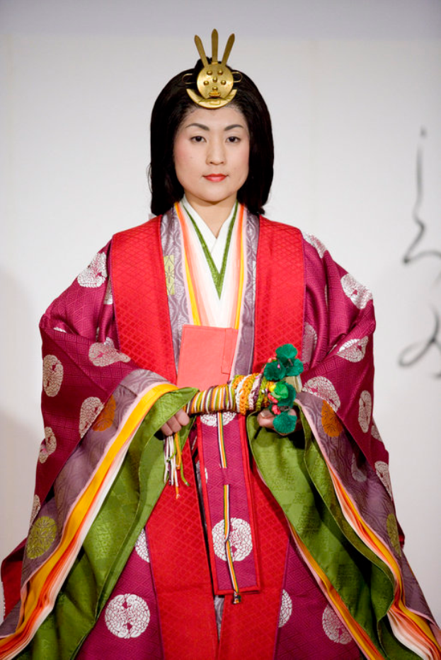 Eine Frau in einem traditionellen japanischen Kimono, mit einem leuchtend roten und grünen Kleid mit aufwändigen Mustern, steht selbstbewusst auf einem Laufsteg, ihr Haar ist ordentlich zu einem Knoten gebunden.