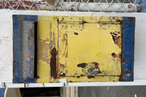 Ein gelb-blauer Metalltresor steht auf einem weißen Wall, mit einem Metallzaun im Hintergrund.