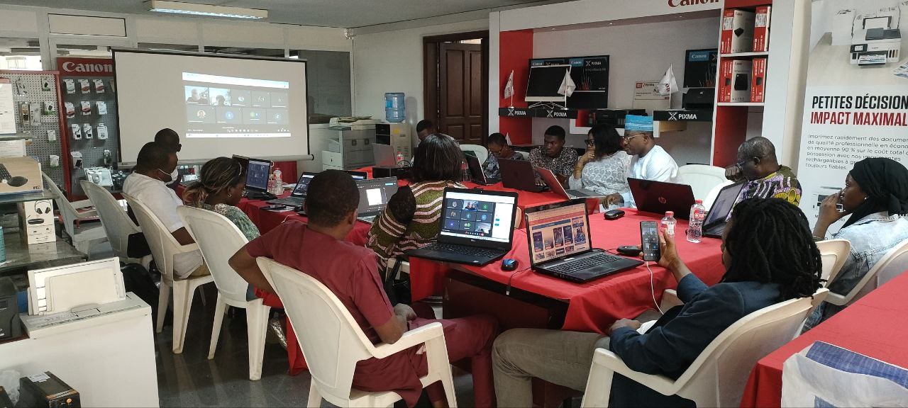 Gruppe von Menschen um einen Tisch mit Laptops, Flaschen und anderen Gegenständen während eines digitalen Marketing-Workshops, mit einem Banner, einem Bildschirm, einer Tür und Deckenleuchten im Hintergrund.