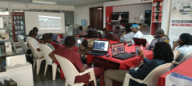 Gruppe von Menschen um einen Tisch mit Laptops, Flaschen und anderen Gegenständen während eines digitalen Marketing-Workshops, mit einem Banner, einem Bildschirm, einer Tür und Deckenleuchten im Hintergrund.