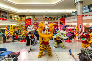 Lebendiges chinesisches Neujahrsfest in einem Einkaufszentrum mit Menschen in Kostümen, die tanzen, und anderen, die auf Stühlen sitzen und mobile Telefone halten, umgeben von Fahnen, Bannern, Dekorationen und Lichtern.