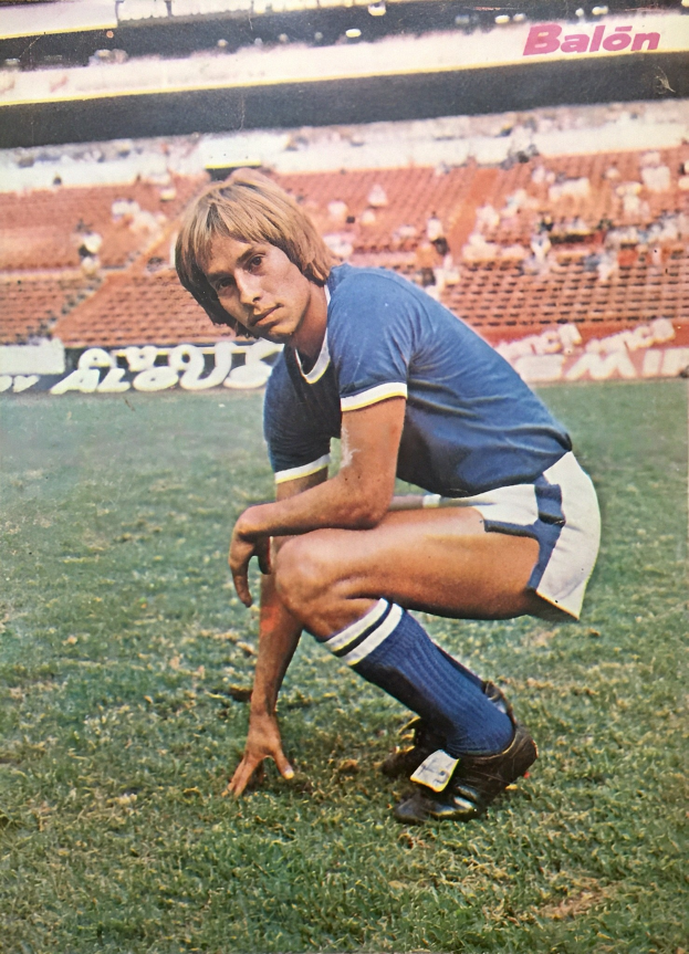 Ein Mann kniend auf einem Fußballfeld umgeben von Menschen auf Stühlen und Tafeln mit Text.