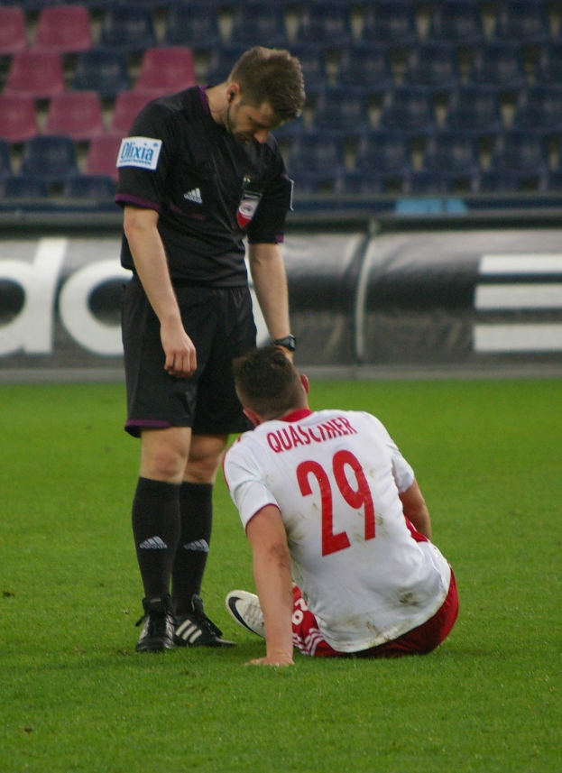 Ein Fussballspieler In Sportbekleidung Sitzt Auf Dem Boden Neben Einem Schiedsrichter In Einer Stadionatmosphäre, Mit Tafeln Und Stühlen Im Hintergrund.