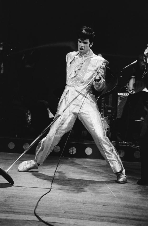 Elvis Presley performs on stage at the Apollo Theater in Las Vegas, wearing a white jumpsuit and holding a microphone, with another musician to his right and stage instruments and lights in the background.