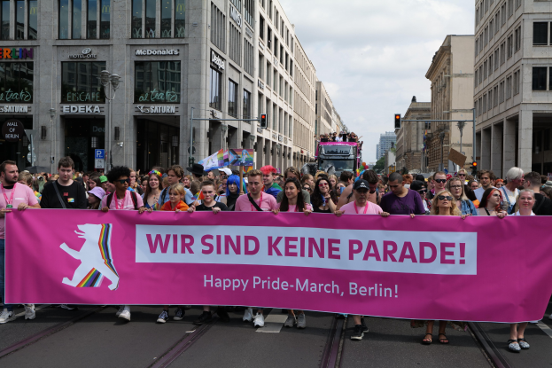 Eine Gruppe von Menschen marschiert auf einer Straße in Berlin und hält eine rosa Schriftzug 'Happy Pride March' hoch, mit Gebäuden, Laternenpfählen und Verkehrszeichen in der Straße unter einem bewölkten Himmel.