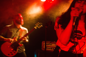 Eine Frau singt in ein Mikrofon auf der Bühne, während ein Mann im Hintergrund eine akustische Gitarre spielt, mit Deckenleuchten, die die Szenerie beleuchten.