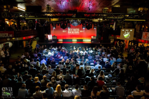 Eine große Menschenmenge, die auf Stühlen vor einer Bühne sitzt, die von Scheinwerfern beleuchtet und von Absperrungen umgeben ist, mit Bildschirmen und Bannern, auf denen Text steht, und einem Wasserzeichen in der linken unteren Ecke, das eine Atmosphäre der Aufregung und Vorfreude für ein Online-Marketing-Rockstars-Event schafft.