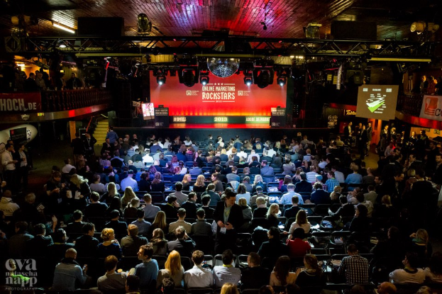 Eine große Menschenmenge, die auf Stühlen vor einer Bühne sitzt, die von Scheinwerfern beleuchtet und von Absperrungen umgeben ist, mit Bildschirmen und Bannern, auf denen Text steht, und einem Wasserzeichen in der linken unteren Ecke, das eine Atmosphäre der Aufregung und Vorfreude für ein Online-Marketing-Rockstars-Event schafft.