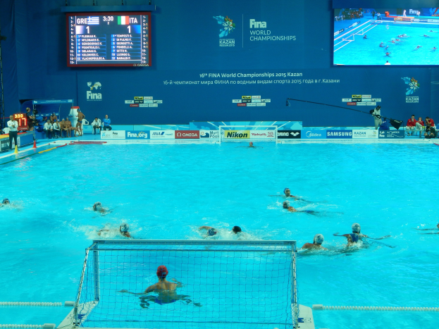 Wasserballspieler bei einem Pool-Wettkampf während der FINA-Weltmeisterschaften 2016 in Kazan, mit Zuschauern links und einem Scoreboard mit Event-Branding im Hintergrund.