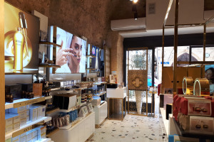 Innenansicht eines Kosmetikladens mit verschiedenen Produkten auf Gestellen, wandmontierten Bildschirmen, Deckenleuchten, einer Klimaanlage und Glastüren, die eine Person, ein Auto und ein Gebäude zeigen.