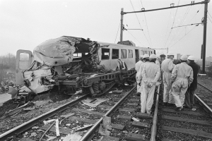 Ein Schwarz-Weiß-Bild einer Zugkollision mit Menschen in Helmen, die drumherum stehen, Strommasten, Bäumen, Gebäuden und einem klaren Himmel im Hintergrund.