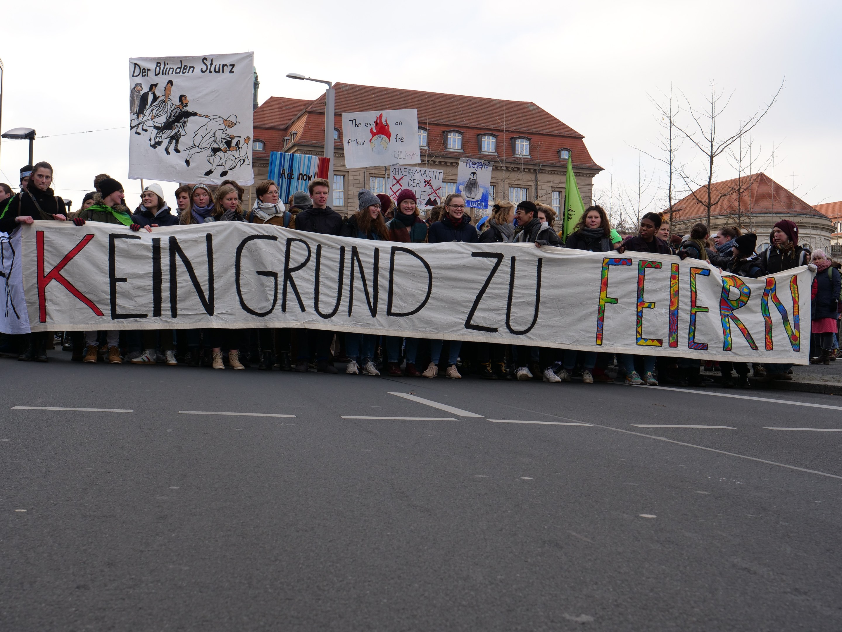 Gruppe von Menschen mit einem Banner "Kein Grund zu Feiern" im Protest gegen die Sparpolitik der deutschen Regierung, mit Straßeninfrastruktur und Gebäuden im Hintergrund bei klarem Himmel.