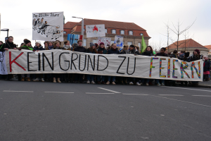 Gruppe von Menschen mit einem Banner "Kein Grund zu Feiern" im Protest gegen die Sparpolitik der deutschen Regierung, mit Straßeninfrastruktur und Gebäuden im Hintergrund bei klarem Himmel.