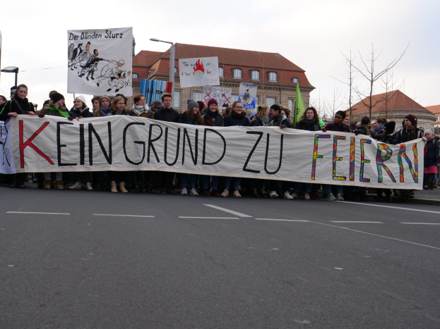 Gruppe von Menschen mit einem Banner "Kein Grund zu Feiern" im Protest gegen die Sparpolitik der deutschen Regierung, mit Straßeninfrastruktur und Gebäuden im Hintergrund bei klarem Himmel.