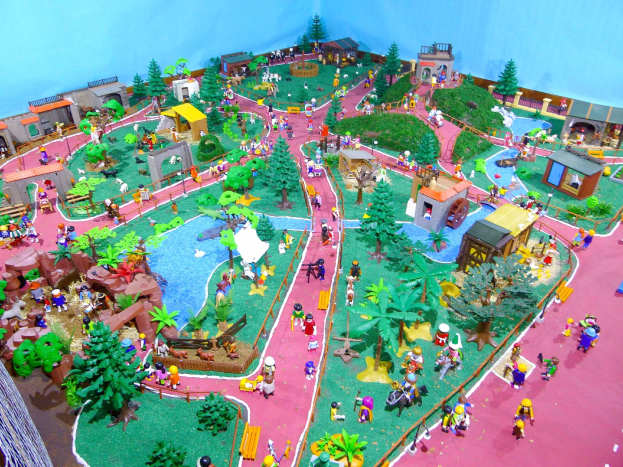 Ein Miniatur-Modell eines Zoos mit vielen Menschen darauf, umgeben von Bäumen, Häusern, Pflanzen, Gras und einem Zaun, mit einer Wand im Hintergrund und Text unten.