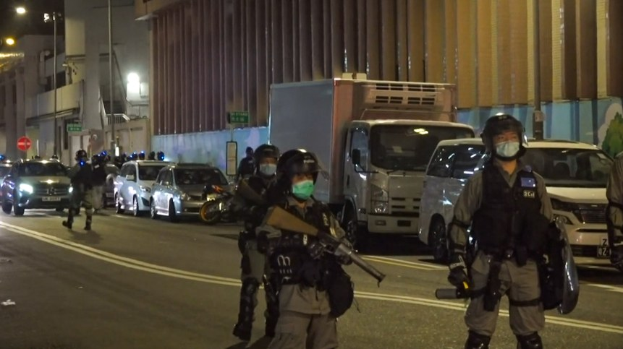 Eine Gruppe von Polizisten in Helmen, Handschuhen und Gesichtsmasken steht auf einer Straße in der Nacht, einige halten Waffen, mit Fahrzeugen, Laternenmasten, Schildern und Gebäuden im Hintergrund.