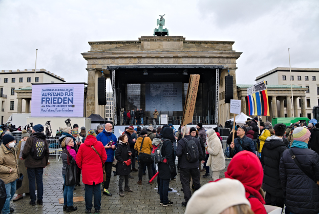 Eine Menschenmenge steht vor einem Gebäude mit einer Bühne mit Rednern und einem Bildschirm, umgeben von Fahnen und Transparenten mit Text, in Berlin während einer Demonstration.
