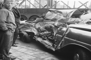 Schwarz-weißes Bild eines verbeulten Autos am Straßenrand mit mehreren Menschen in der Nähe, einem Lastwagen und einer Brücke im Hintergrund.