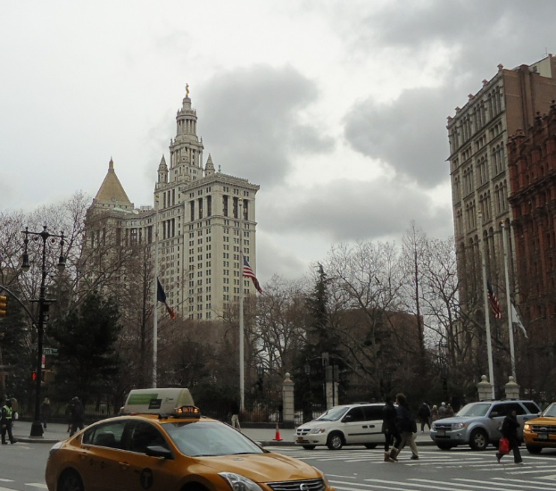 Ein gelbes Taxi fährt auf einer von Hochhäusern gesäumten Stadtstraße, Fußgänger auf dem Gehweg, Verkehrskegel, Laternen, Fahnen mit Stangen und Bäume, unter einem bewölkten Himmel.