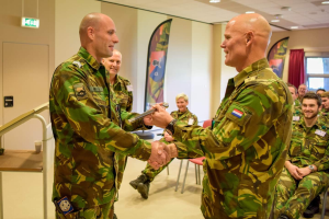 Zwei Männer in Militäruniformen geben sich die Hand vor einer sitzenden Gruppe, mit einem Tisch, Geländer, Fahnen, einer Tür, einem Schild, einem Fenster mit Vorhang und Deckenleuchten im Hintergrund, was auf eine deutsche Armee-Versammlung hinweist.