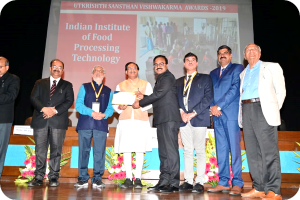 Eine Gruppe von Männern auf einer Bühne mit einem Zertifikat, im Hintergrund ein Bildschirm mit der Aufschrift "Indian Institute of Food Processing Technology Awards 2019" und Blumenschmuck.