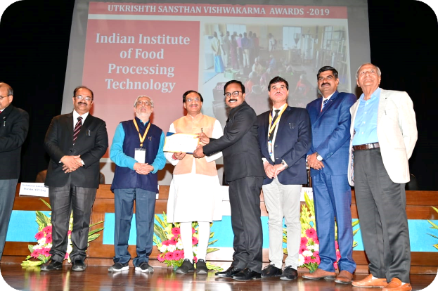 Eine Gruppe von Männern auf einer Bühne mit einem Zertifikat, im Hintergrund ein Bildschirm mit der Aufschrift "Indian Institute of Food Processing Technology Awards 2019" und Blumenschmuck.