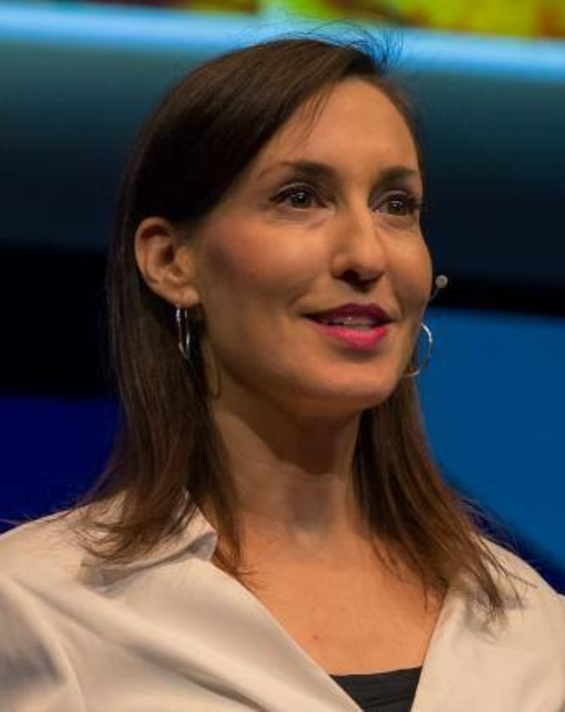 Frau in weißer Jacke und schwarzem Oberteil, die vor einem blauen und gelben Hintergrund sitzt.