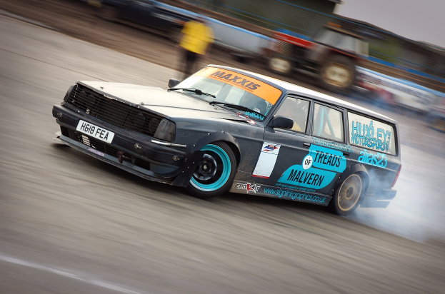 Ein Auto auf einer Rennstrecke, das Rauch ausstößt, mit anderen Fahrzeugen und Zuschauern im Hintergrund.
