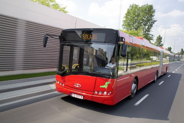 Ein roter Bus fährt auf einer Straße neben einem Gebäude, mit einer Person darin sichtbar.