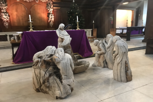 Krippe in einer Kirche mit Statuen, einem Tisch mit einer Decke und Kerzen, Stühlen, einem geschmückten Weihnachtsbaum und einer Wand im Hintergrund.