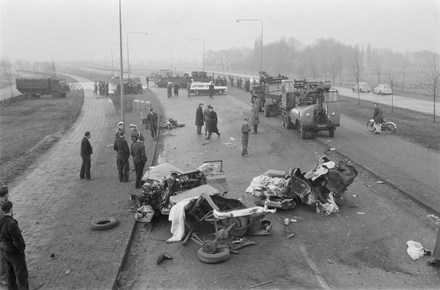 Schwarze und weiße Szene eines Autounfalls am Straßenrand mit mehreren Fahrzeugen und einer Gruppe von Menschen drumherum, Laternenpfähle, Bäume und den Himmel im Hintergrund.