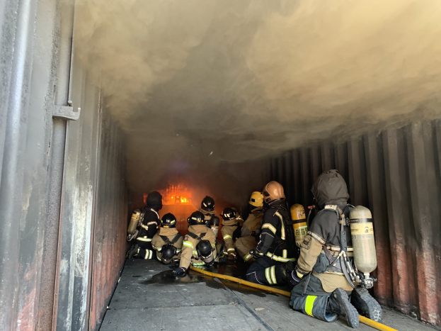 Feuerwehrleute in Schutzausrüstung arbeiten in einem raucherfüllten Tunnel, einer hält einen Schlauch, mit Flammen im Hintergrund und einer Wand auf der linken Seite.