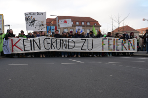 Demonstranten mit einem Banner "Kein Grund zu Feiern" gegen deutsche Sparmaßnahmen, mit Gebäuden, Bäumen und einem klaren Himmel im Hintergrund.