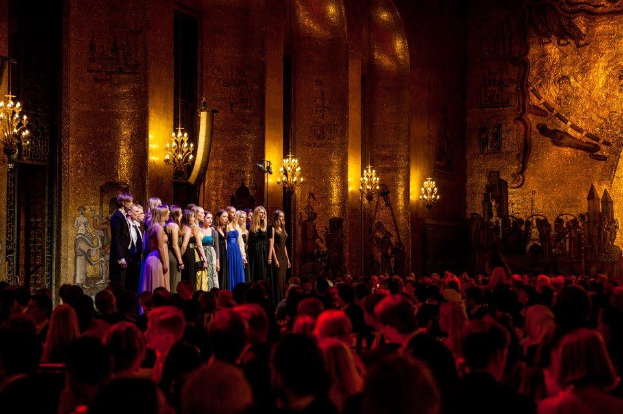 Eine Gruppe von Menschen auf einer beleuchteten Bühne im Royal Opera House in London, mit Zuschauern darunter.